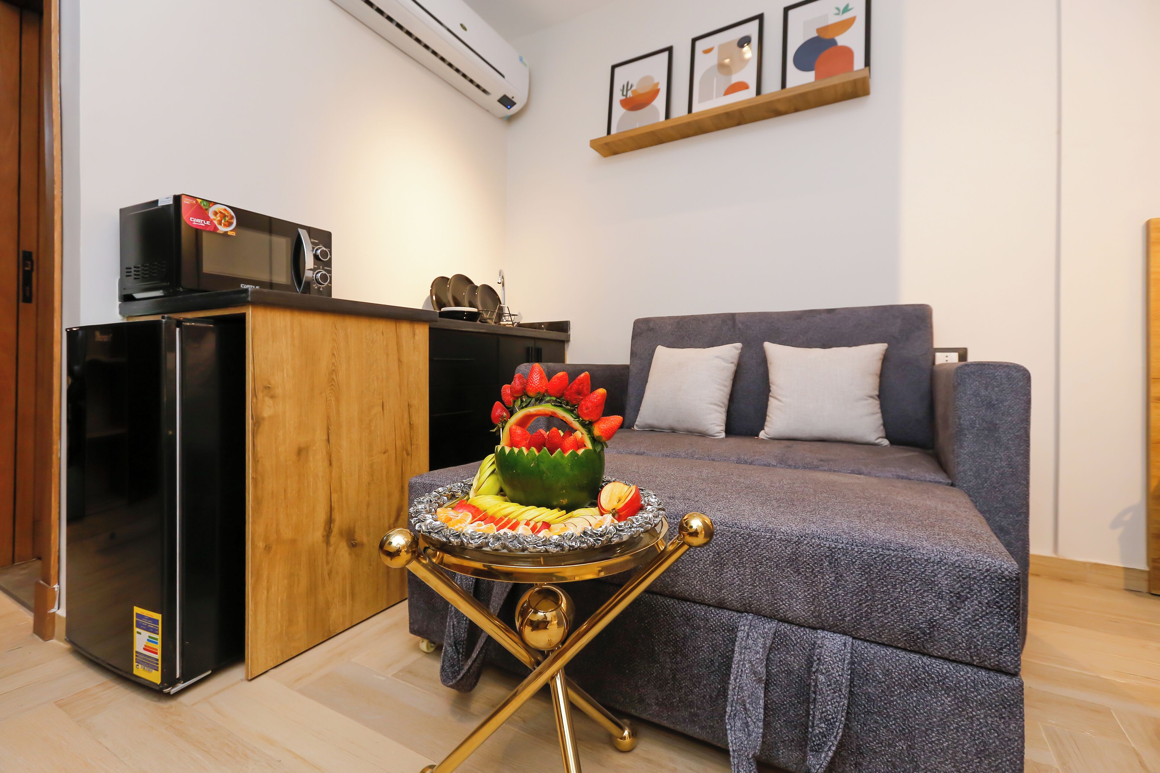 A compact living space with a kitchenette, a grey sofa, and a decorative fruit arrangement on a small table.