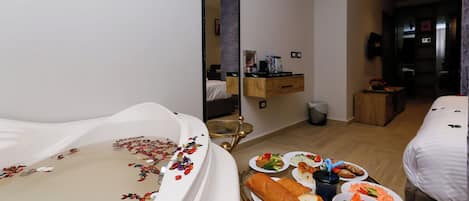 A hotel room with a large bathtub, a dining table with plates of food, a mirror, and a wooden cabinet.