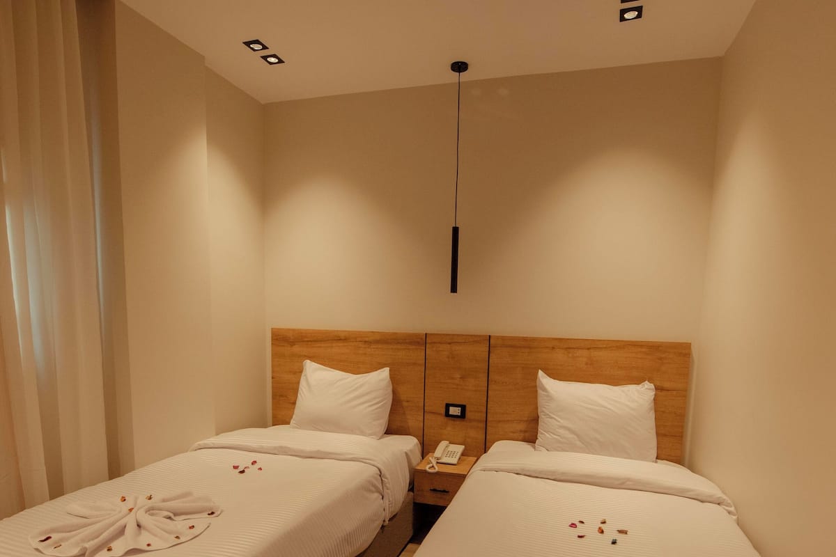 A hotel room with two single beds, each with a wooden headboard, white linens, and a small bedside table with a lamp.