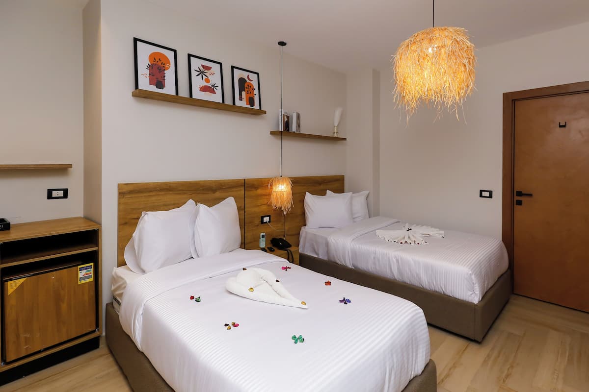 A hotel room with two beds, a wooden wardrobe, a bedside table with a lamp, and a door.