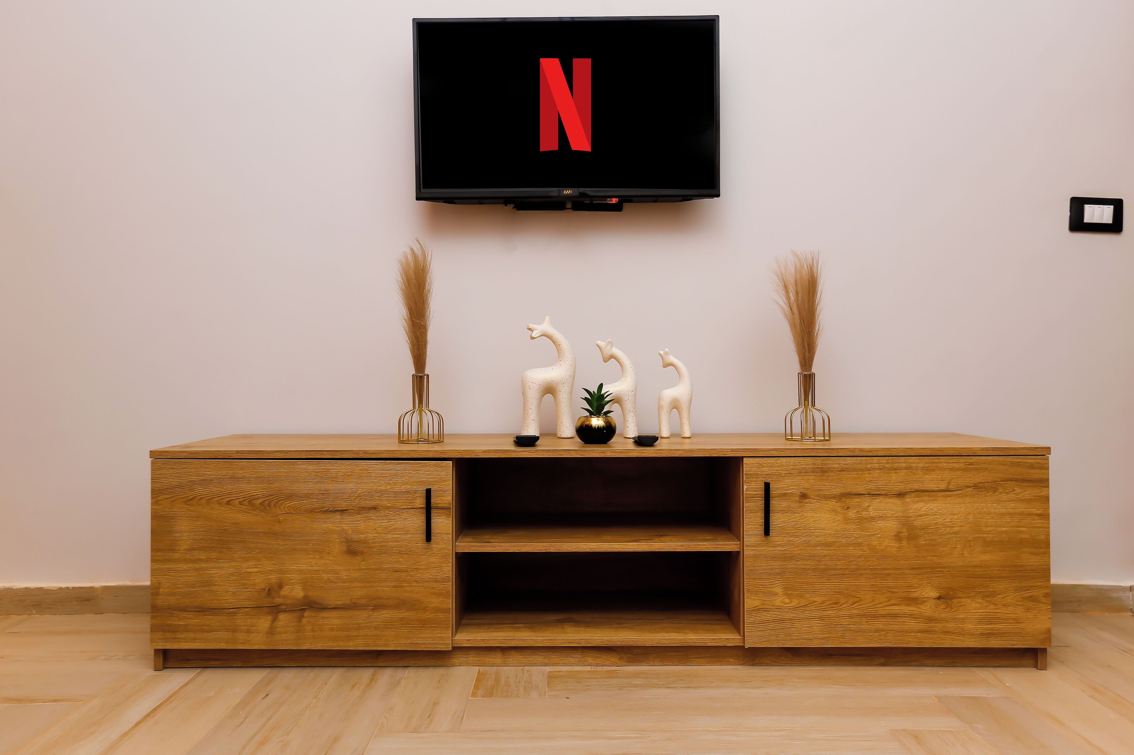 Un mueble de televisión de madera con un televisor de pantalla plana mostrando el logotipo de Netflix, dos jarrones decorativos y dos figuritas de animales encima.