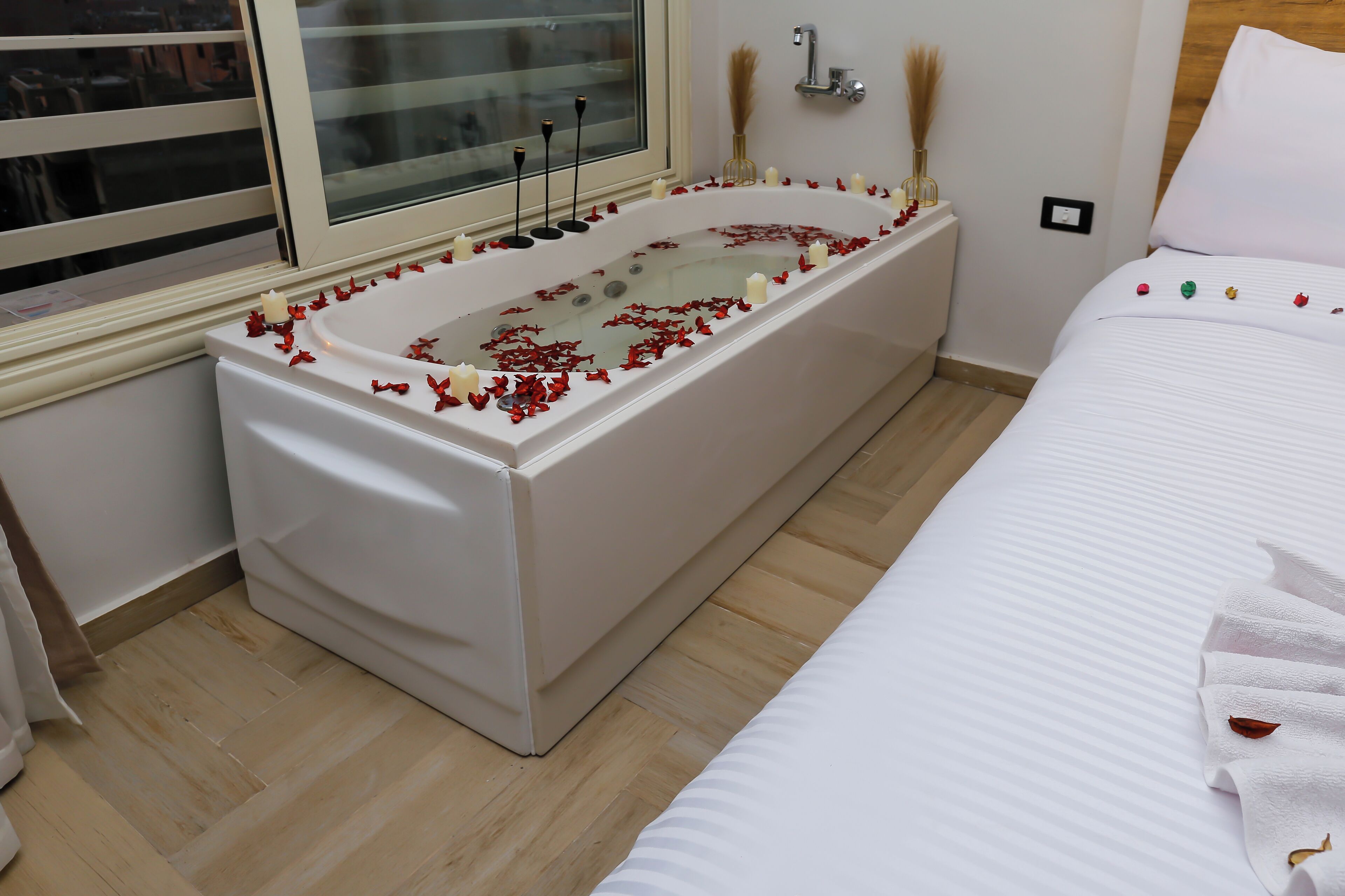 A modern bathroom with a freestanding bathtub, a window with blinds, a wooden floor, and a bed with a white bedspread and a towel.