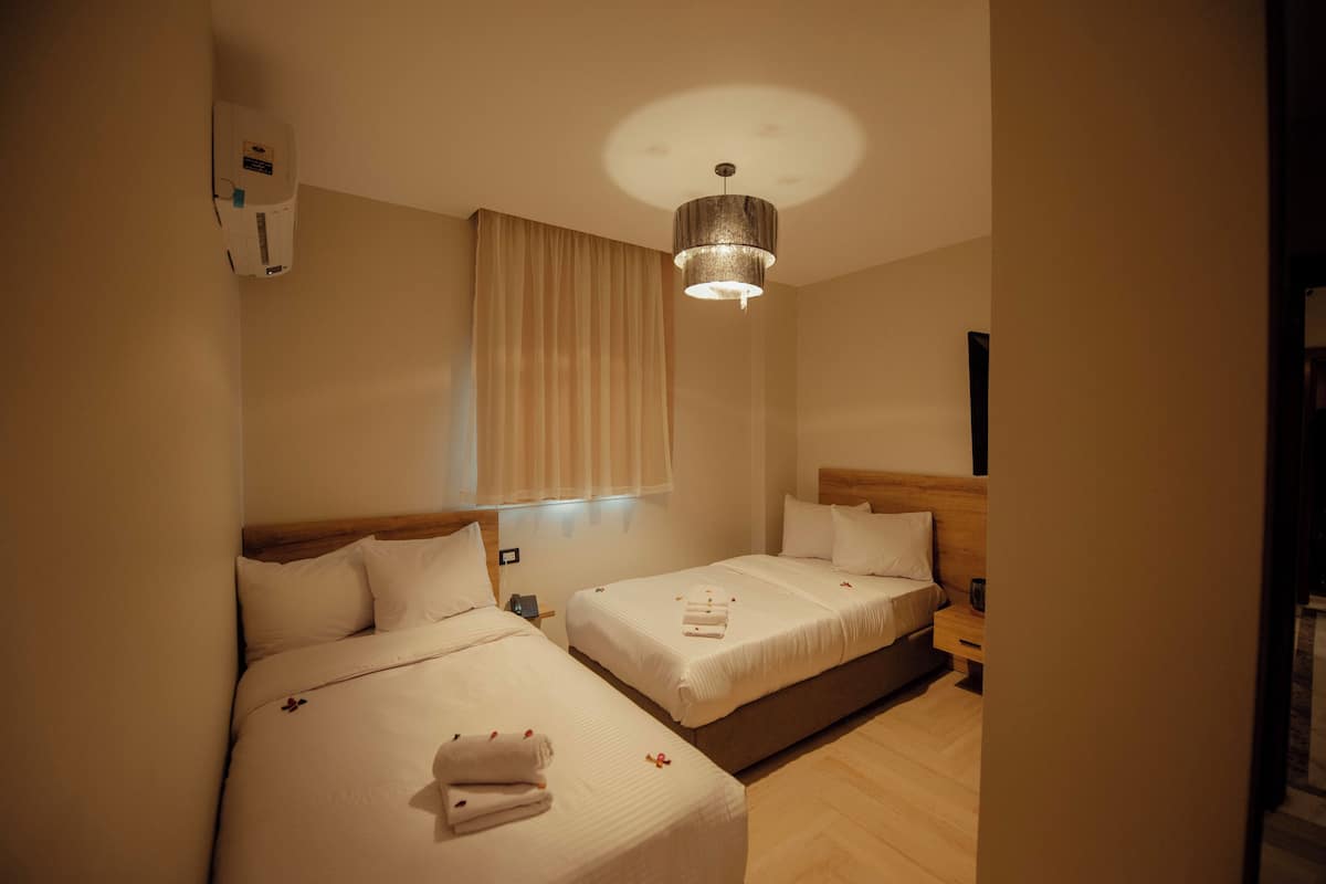 A hotel room with two beds, a ceiling light, a window with curtains, and a wall-mounted air conditioner.