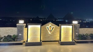 Ein Nachtblick auf einen Gebäudeeingang mit beleuchteten Säulen und einer zentralen sternförmigen Leuchte, vor der Pyramiden zu sehen sind.