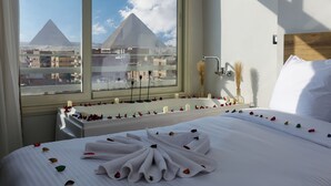 Ein Hotelzimmer mit Bett, einem Fenster mit Blick auf Pyramiden, einer Badewanne mit Rosenblüten und einem Handtuchhalter.