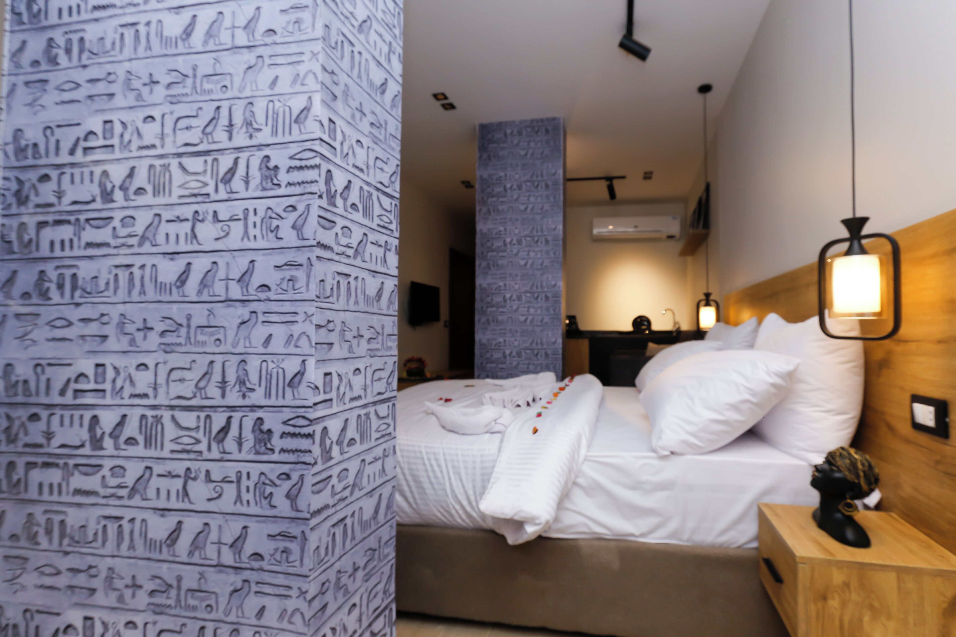 Una habitación de hotel moderna con una cama, una mesita de noche de madera, una pared con jeroglíficos y una lámpara colgante.