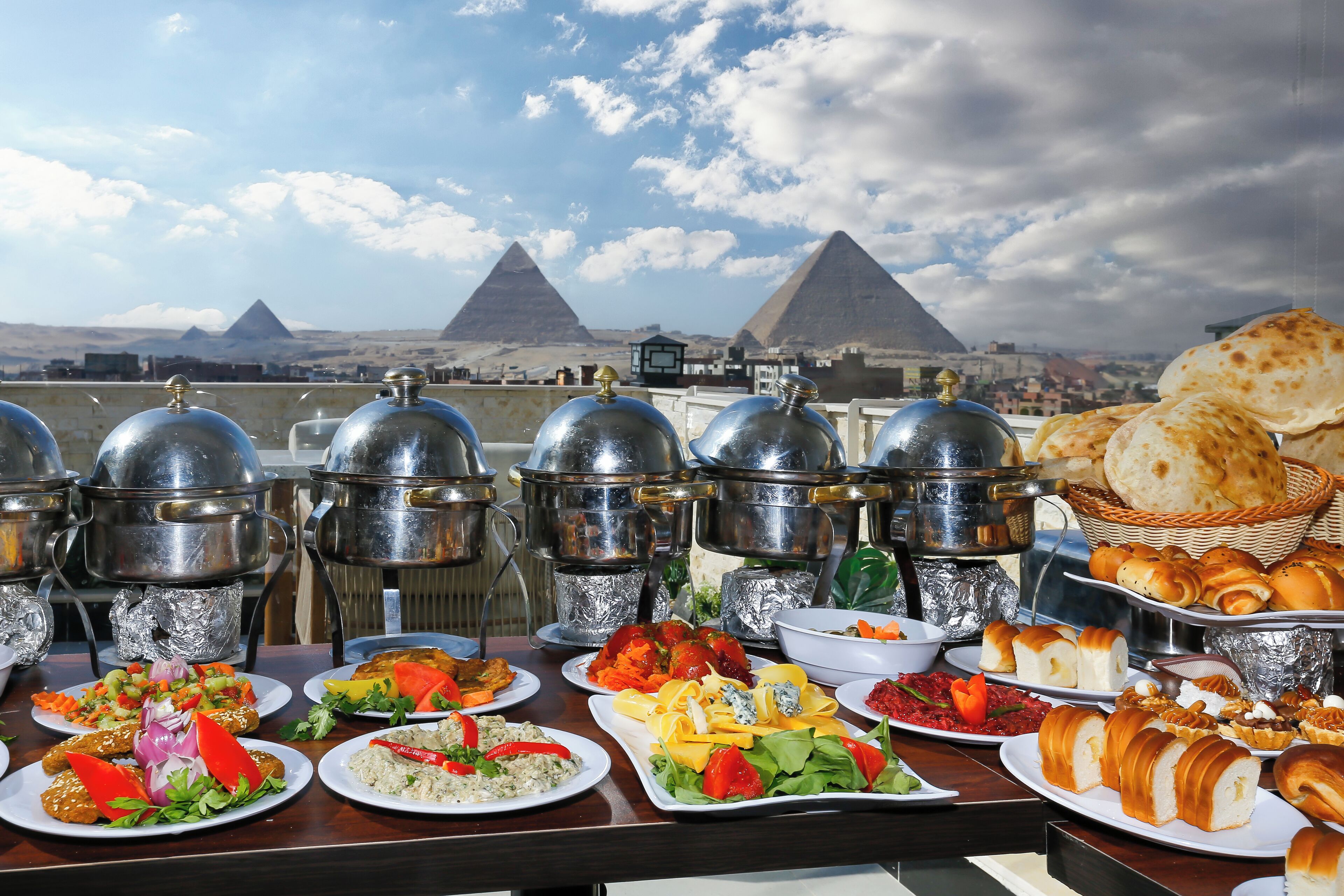 Una mesa de buffet con diversos platos y pan, con pirámides y un cielo nublado al fondo.