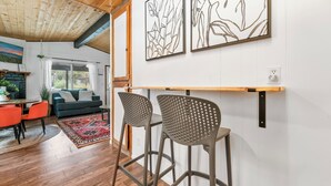 Una sala de estar moderna con techo de madera, un sofá, una mesa de centro y dos taburetes de bar. Se observan obras de arte enmarcadas en la pared y una ventana con cortinas.