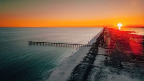 An aerial view of a beach with a pier extending into the water, a road parallel to the beach, and buildings along the coastline.