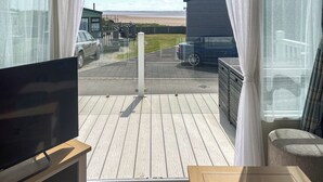 A balcony with a TV, a wooden floor, and a view of a beach and parked cars. - 3 bedroom accommodation in Lancaster (Lancaster)