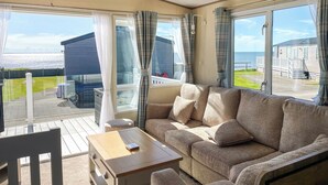 A cozy living room with a sofa and a table, large windows with curtains, and a view of the sea and other buildings. - 3 bedroom accommodation in Lancaster (Lancaster)