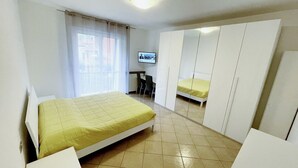 Quarto com uma cama, escrivaninha, cadeira, televisão e guarda-roupa.