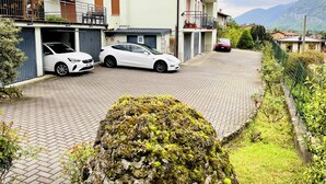 Um bairro residencial com dois carros estacionados, um caminho de acesso pavimentado e uma paisagem verdejante com uma montanha ao fundo.