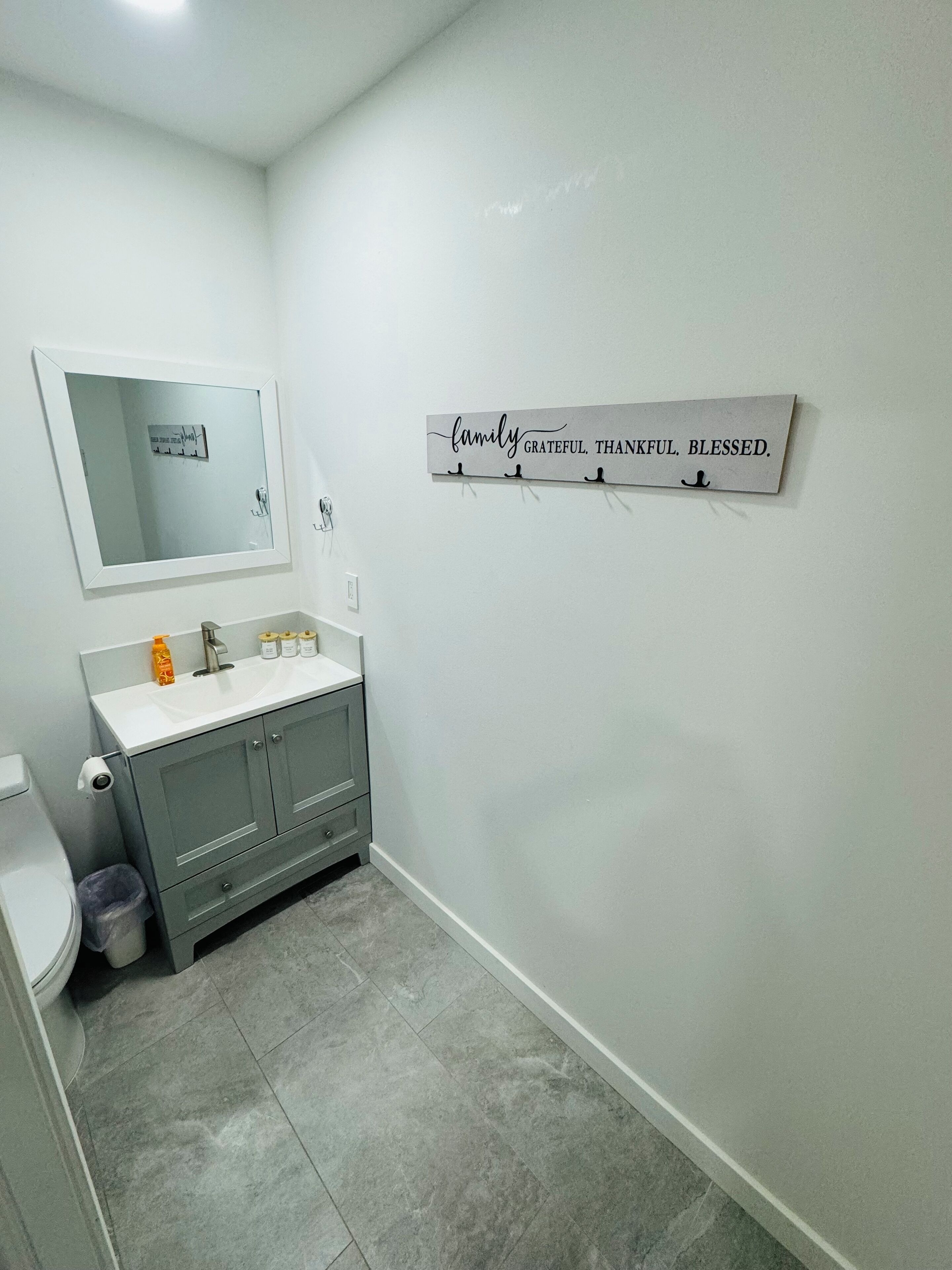 A bathroom with a white sink, grey cabinets, and a mirror. There is a wall-mounted sign with the words "family grateful thankful blessed."