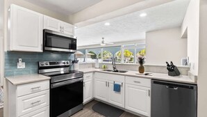 Una cocina moderna con armarios blancos, electrodomésticos de acero inoxidable y un amplio ventanal con vistas a palmeras.