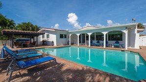 Un área junto a la piscina con sillas de descanso, un espacio de estar cubierto y una piscina de color azul cristalino.