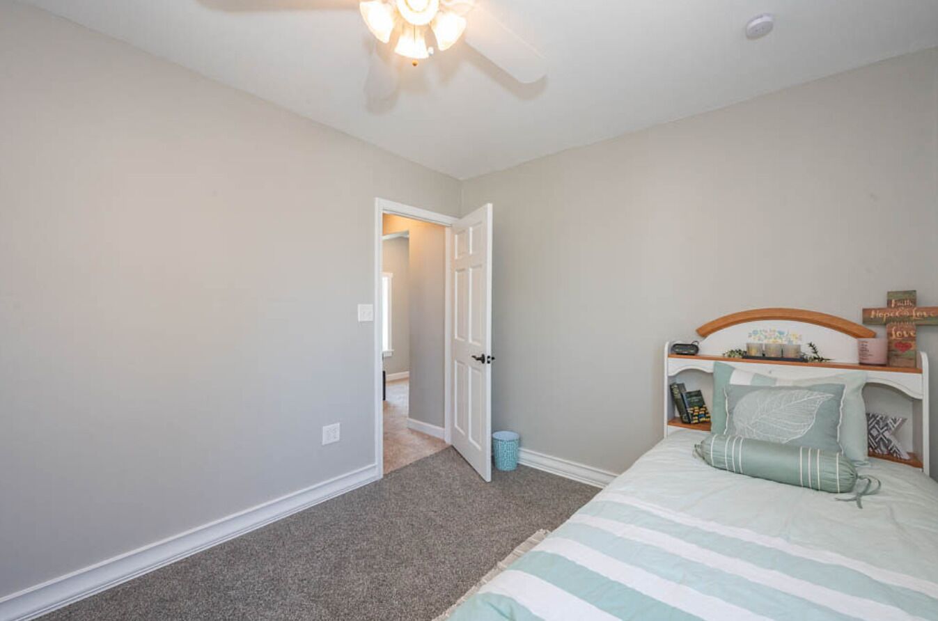 Une chambre à coucher comprenant un lit, une table de chevet, un ventilateur de plafond et une porte donnant sur une autre pièce.