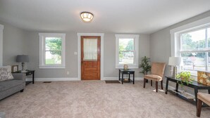 A spacious living room with a carpeted floor, a wooden door, two windows, a sofa, a chair, a small table, and a lamp. - Beautiful 3 Bedroom Downtown Salem Home (Salem)