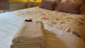 Un lit bien fait, recouvert d’une courtepointe à motifs floraux, avec des oreillers et une serviette pliée, sur laquelle est posé un cœur rouge.