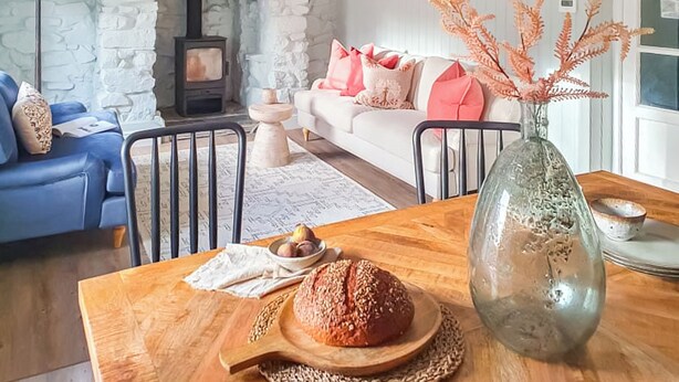 A cozy living room with a stone fireplace, a blue sofa, a wooden dining table with a loaf of bread, and a vase with dried plants. - 2 bedroom accommodation in Bodmin (Bodmin)