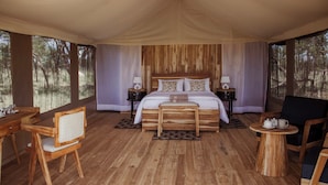 A tented room with a wooden bed, two nightstands, a wooden chair, a round wooden table, and a view of trees. - Conserve Safari Serengeti (Serengeti)