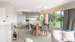 Ein modernes Wohnzimmer mit Essbereich, einer Küche im Hintergrund und großen Fenstern mit Vorhängen.