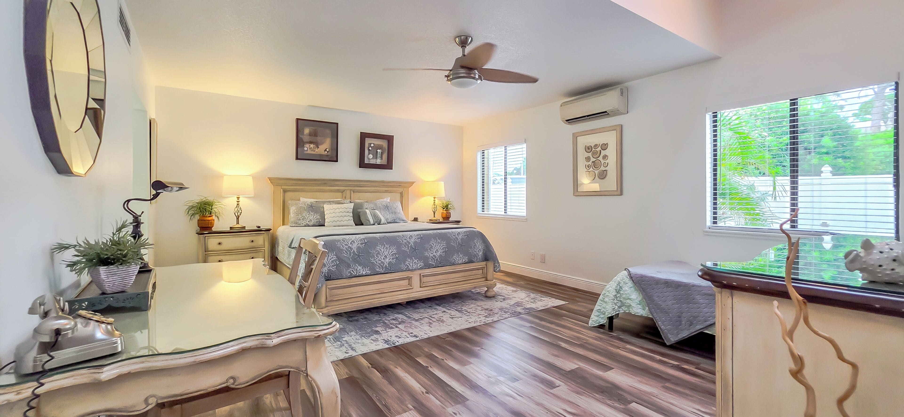 A bedroom with a bed, bedside tables, a desk, a chair, a ceiling fan, and a window with blinds.