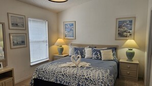 A bedroom with a large bed, two bedside tables, a window with blinds, and framed pictures on the walls.