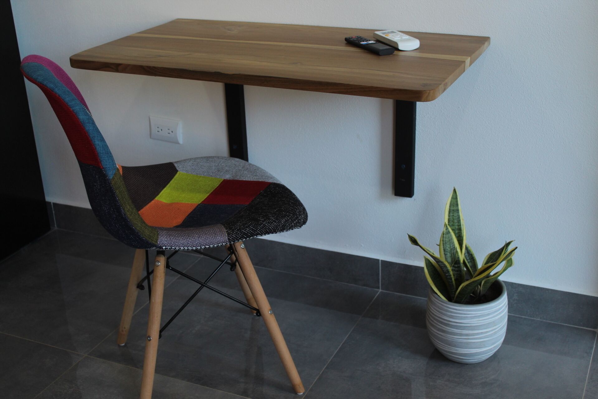 Une chaise avec un siège aux motifs colorés, une table en bois avec une télécommande et une plante en pot.