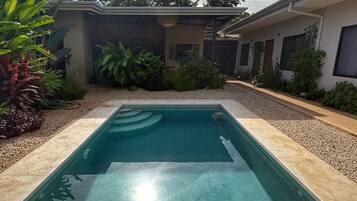 A swimming pool with a tiled deck, surrounded by a garden with various plants and a building in the background.