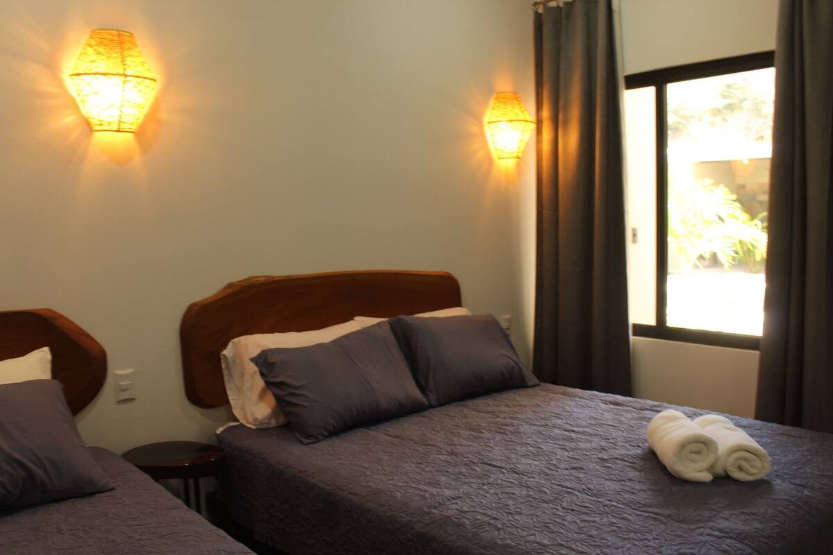 A hotel room with two beds, each with a headboard, a window with curtains, and a view of greenery outside.