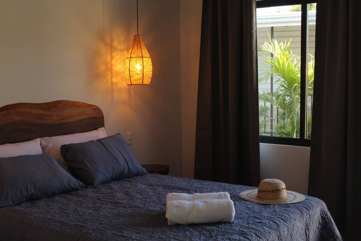 A bedroom with a bed, a wall-mounted light, a window with curtains, and a hat on the bed.