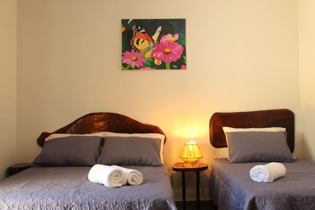A hotel room with two beds, each with a rolled towel, a lamp on a side table, and a painting of a butterfly on a flower on the wall.