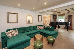 Een groene hoekbank met decoratieve kussens, een rond gouden bijzettafeltje en twee groene poefjes in een kamer met een piano en een stenen muur.