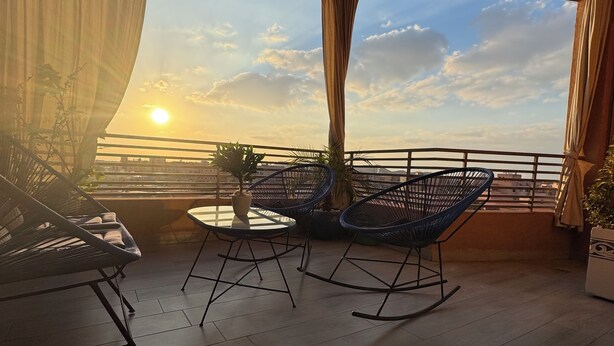Un balcón con vistas a la puesta de sol, que incluye una mesa, dos sillas mecedoras y una planta en maceta.