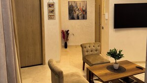 Una sala de estar moderna con sofá, sillón y mesa de centro. Hay un cuadro en la pared, una lámpara de techo y un televisor de pantalla plana montado en la pared.