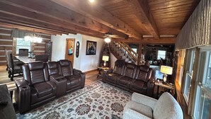 A spacious living room with a large brown leather sofa, a smaller white armchair, and a patterned rug. A staircase with wooden railings leads to an upper level. - Big Kahuna Mauka, Beautiful Mountain View 4-BR Cabin in charming Waynesville (Waynesville)
