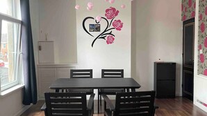 A dining area with a wooden table and chairs, a decorative wall with a heart and roses, and a window with curtains.