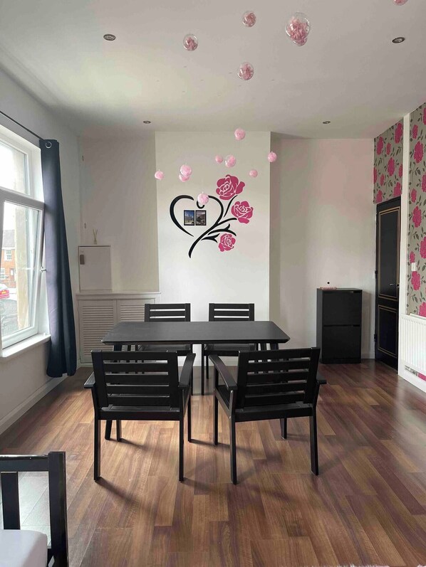 A dining area with a wooden table and chairs, a decorative wall with a heart and roses, and a window with curtains.