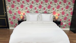 A neatly made bed with white bedding and a black and white striped bottom edge, positioned against a floral wallpaper backdrop.