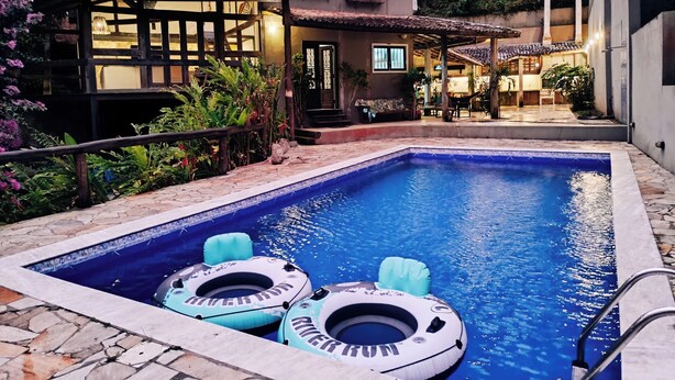 A swimming pool with two inflatable rings, surrounded by a stone deck and a house with a tiled roof and large windows. - Refugio Do Frances Ghesthouse (São Sebastião)