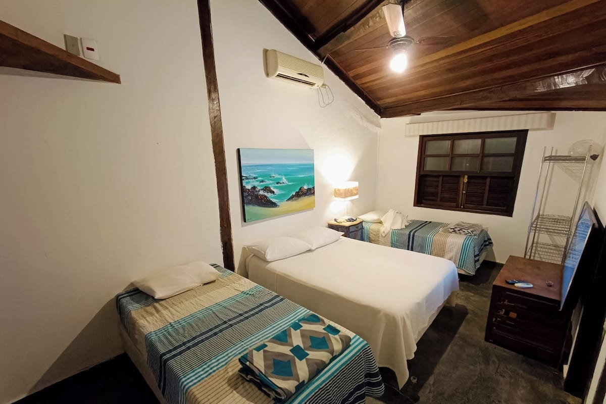 A room with two beds, a painting of a beach, an air conditioner, a lamp, and a window with a view.