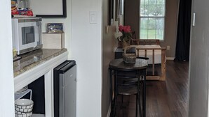 Una cocina con microondas, cafetera y estantes de almacenamiento. Hay una zona de comedor con mesa y sillas, y una ventana con persianas.