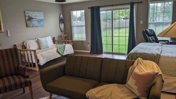 Una habitación acogedora con cama, sofá y sillón. Hay dos ventanas con cortinas, y un cuadro en la pared.