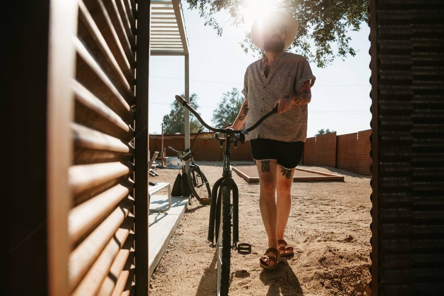 Uma pessoa ao lado de uma bicicleta em área externa coberta, com chão de areia e cerca de metal ao fundo.