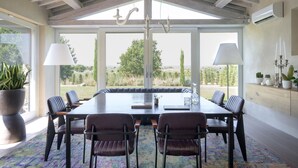 Un comedor con una mesa larga, seis sillas y un banco. Hay dos candelabros, una planta en maceta y un aire acondicionado fijado a la pared.