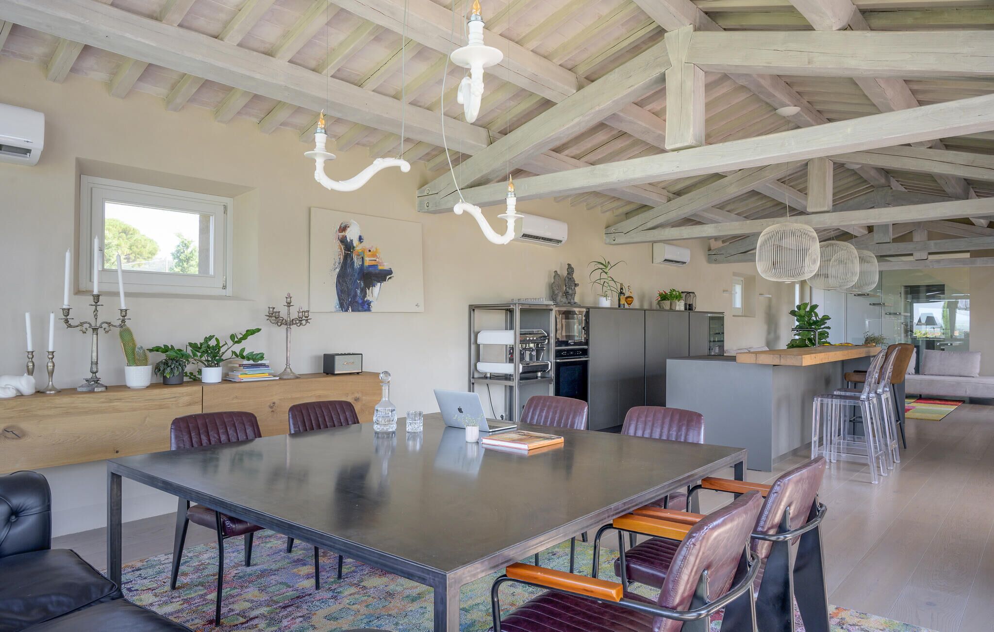 Ein großzügiger Essbereich mit einem langen Tisch und mehreren Stühlen, eine Küche im Hintergrund und eine hohe Decke mit freiliegenden Balken.