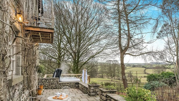 A stone patio with a hot tub, a metal table with chairs, and a view of the countryside. - The B Hive at Buckstone House (Carnforth)
