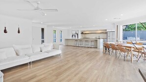 A spacious living area with a white sofa, wooden flooring, a ceiling fan, and a kitchen area with a dining table and chairs. - Black Shack Burleigh (Burleigh Waters)