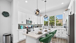 A modern kitchen with white cabinets, a stainless steel refrigerator, and a large island with green bar stools. - Cozy Home Near MedCenter, Uptown, & Downtown (Houston)